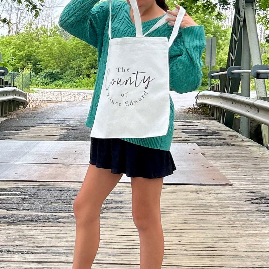 person holding a canvas handbag with "The County of Prince Edward" printed on the bag.